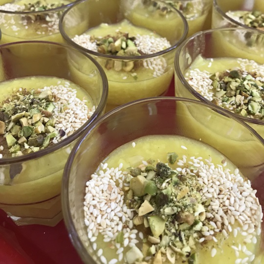 Photo of many small clear glass cups filled with pale yellow rice pudding the top has chopped green pistachios, white sesame seeds, and cinnamon
